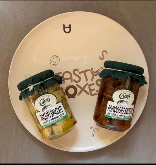 La foto mostra due tipi di sottoli pugliesi su un piatto in ceramica con logo TastyBoxes: carciofi spaccati e pomodori secchi in olio extravergine d'oliva