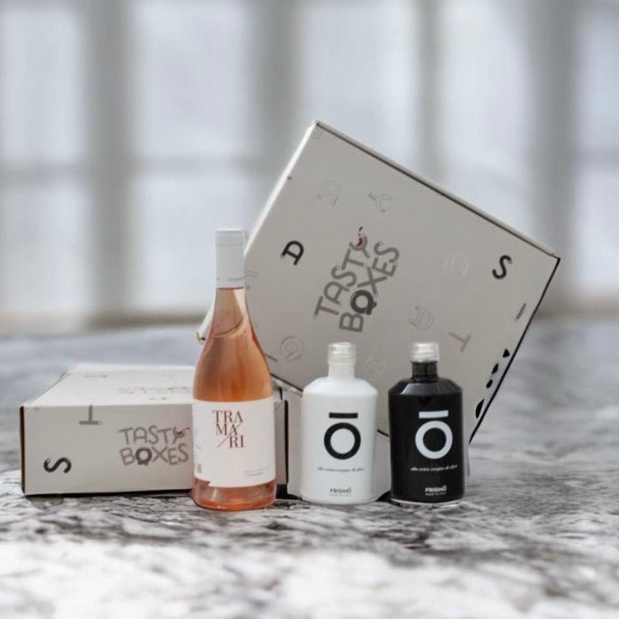 Immagine della scatola con logo Tastyboxes su un tavolo di marmo bianco e grigio, ed i prodotti contenuti nella box: Vino Tramari rosé di primitivo pugliese, confezione bianca olio peranzana Frisino Limited Edition e confezione nera di olio extravergine di oliva coratina Frisino Limited Edition, venduta da TastyBoxes